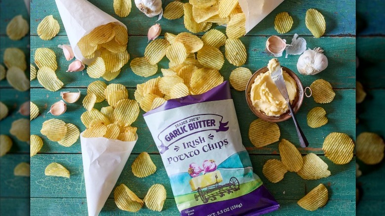 Trader Joe's garlic butter Irish potato chips in bag, scattered around, and in paper sleeves with garlic bulbs and butter on a green wooden table.