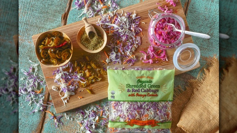 A bag of Trader Joe's organic cole slaw mix in a bag surrounded by spices, jalapenos, pickled cabbage on a wooden cutting board.