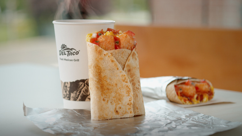 A toasted breakfast wrap on tinfoil with a steaming drink behind it
