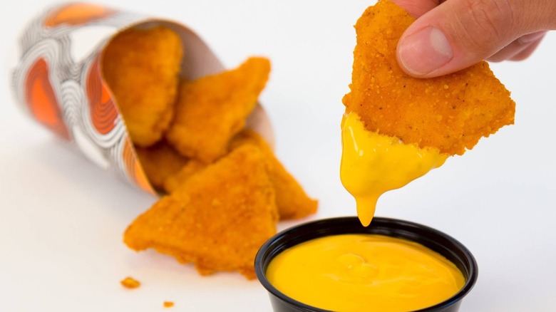 Hand dipping naked chicken chip into nacho sauce with container of naked chicken chips in background