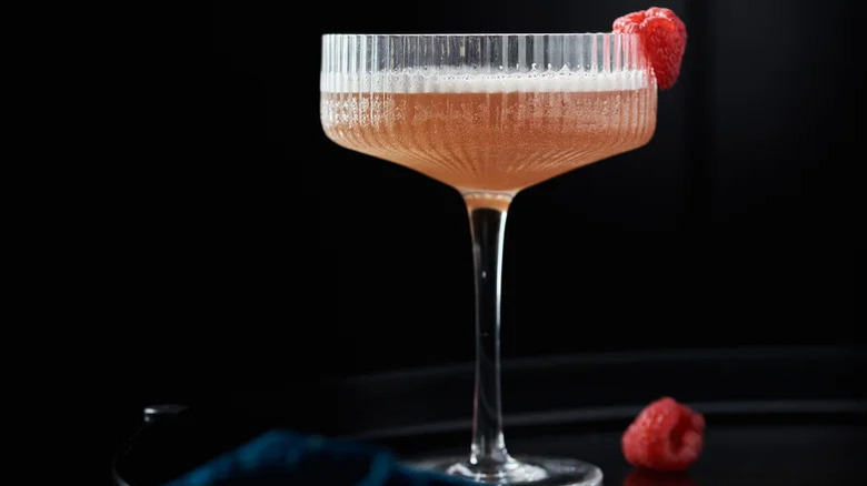 Pink cocktail in coupe glass with raspberry against black background.