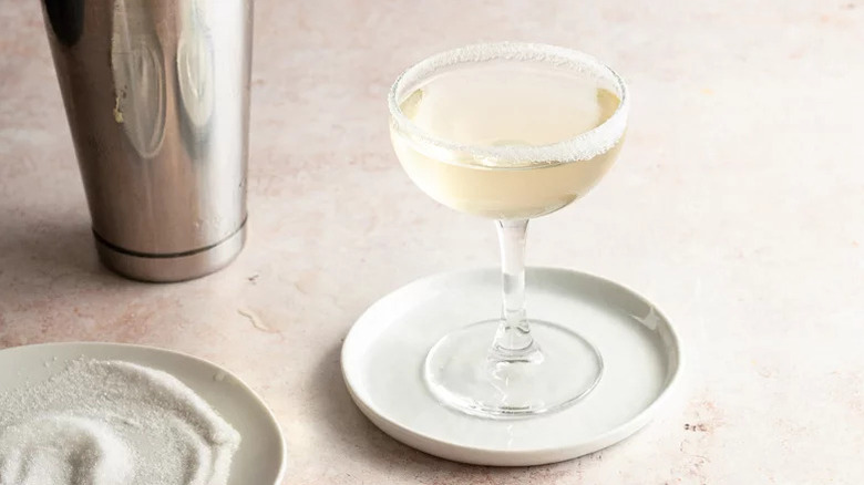 Lemon Drop Martini with sugar rim next to shaker and sugar against white background.