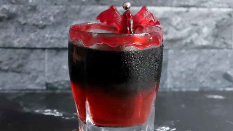 Rocks glass of red and black drink with gummy bat garnish against gray background.