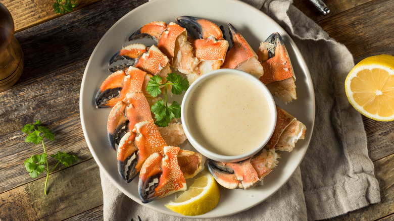 Plate of stone crab claws surrounding small bowl of dipping sauce with lemon wedge sitting on beige cloth on wood surface