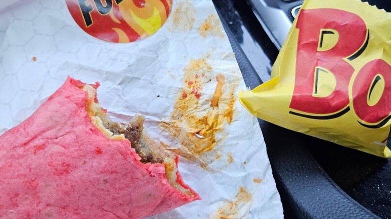 A burrito with a bit missing, made with a red tortilla, sitting in a paper wrapper