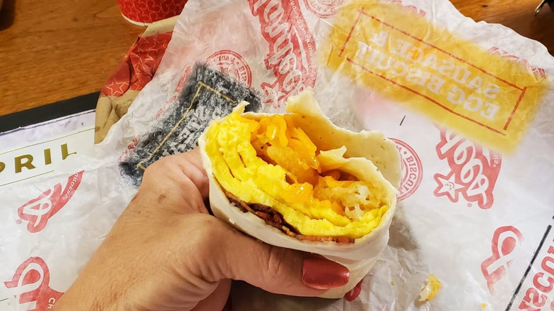 A hand holding a half eaten breakfast burrito full of egg and bacon over a Hardee's wrapper