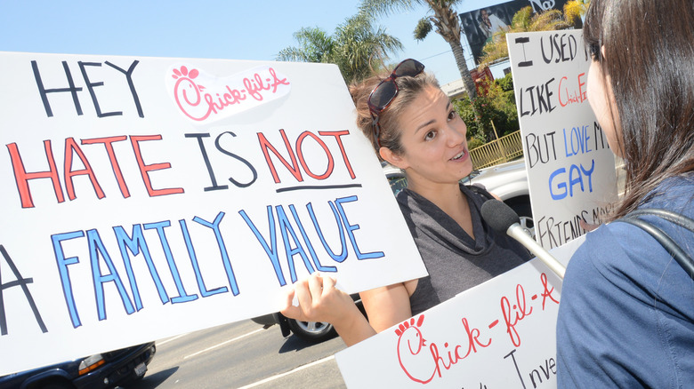 Woman being interviewed holds up a sign that reads "Hey Chick-fil-A, hate is not a family value"