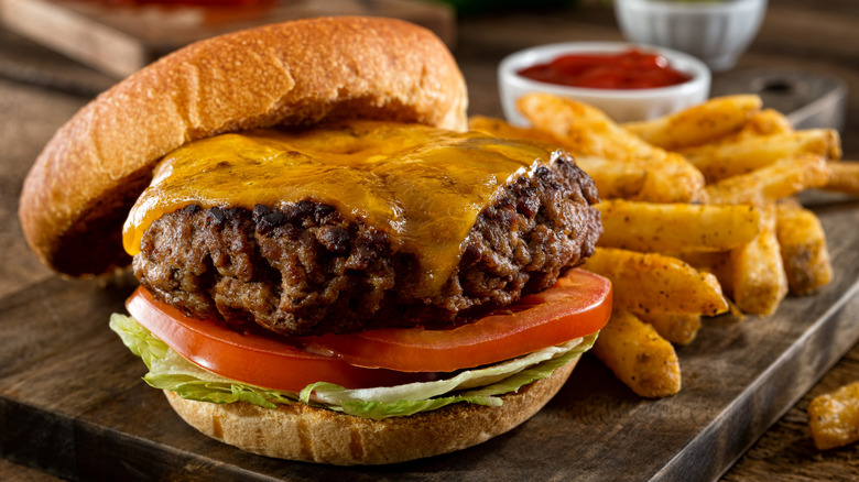 Cheeseburger with top bun taken off next to thick cut french fries and cup of ketchup on dark wooden board