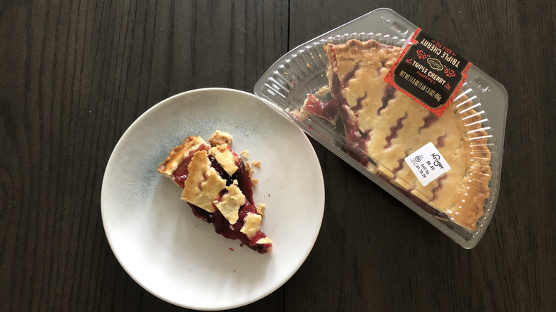 Half of a Kroger's Private Selection Triple Cherry Pie on a wooden table, next a slice on a white plate to the left