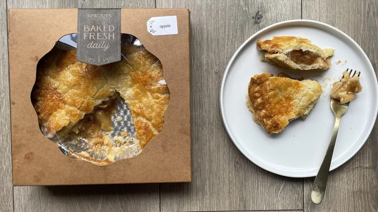 A Sprouts apple pie sits in a box with one slice missing. That slice sits on a white plate on a wooden table