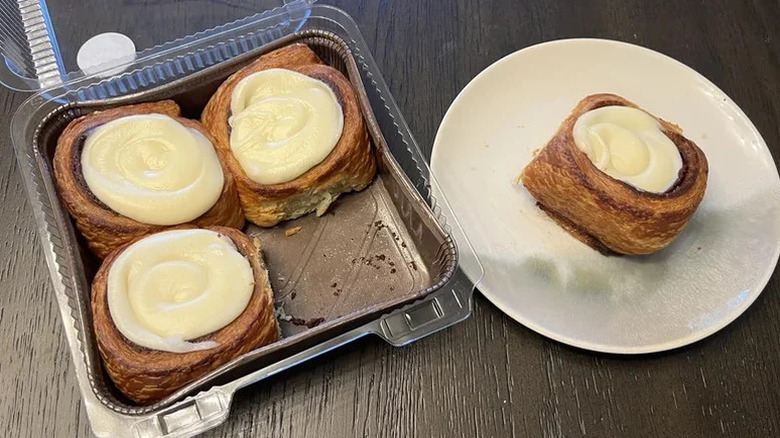 A box of cinnamon buns on a wooden table, next to one bun on a white plate