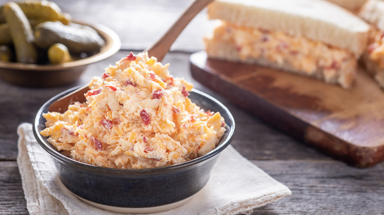 Pimento cheese spread in a bowl, and on a sandwich with a bowl of pickles in background.