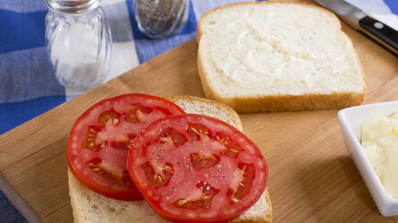 Slices of tomato with mayonnaise, salt, and pepper on white bread, open-faced on wooden cutting board.