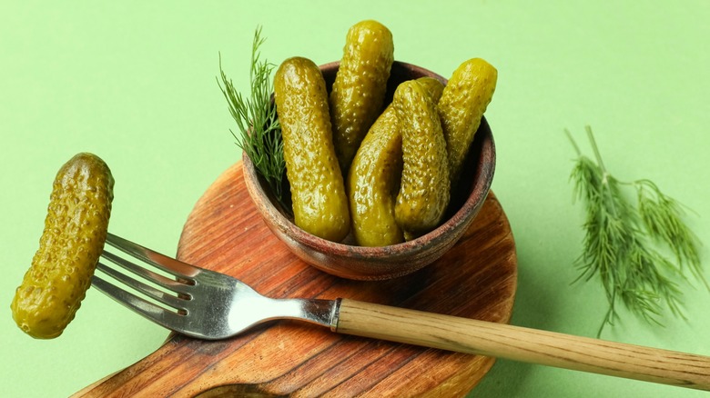 Small brown bowl of pickles on wooden board next to fork with pickle on the sharp end