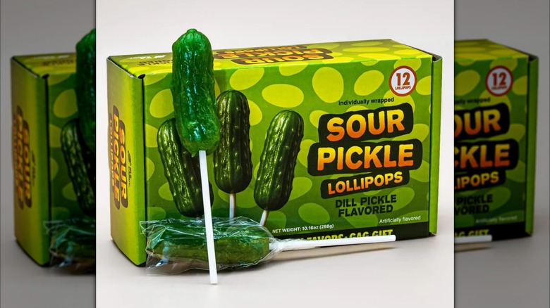 Box of Sour Pickle Lollipops next to two whole lollipops on white surface