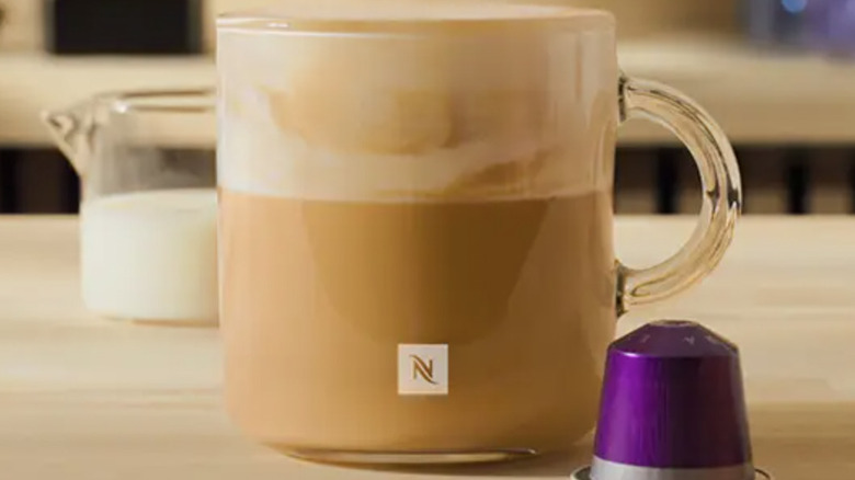 A glass mug of milky coffee and a purple Nespresso pod on a wooden table