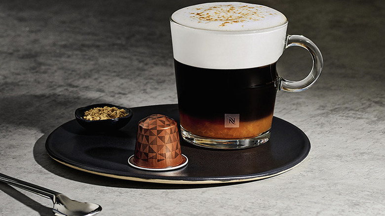 A frothy coffee, a Nespresso capsule, and sugar on a tray beside a spoon