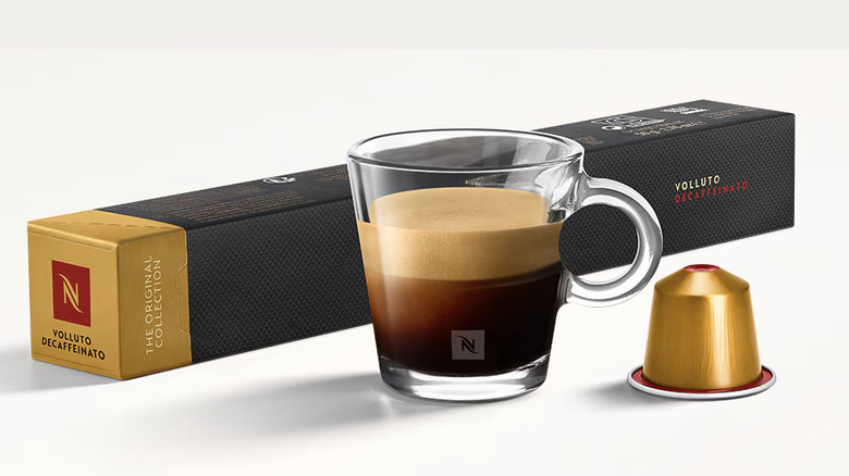 A coffee in a clear mug in front of a pack of Nespresso pods and beside a single gold pod