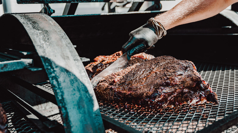 Hand holding knife slicing into full smoked brisket while it's still in the smoker.