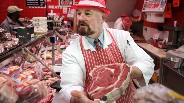 Butcher holding up large raw cut of beef behind counter while customers look on.