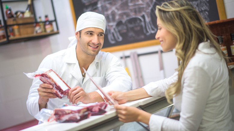 Butcher behind counter holding package of meat with customer in front of counter.