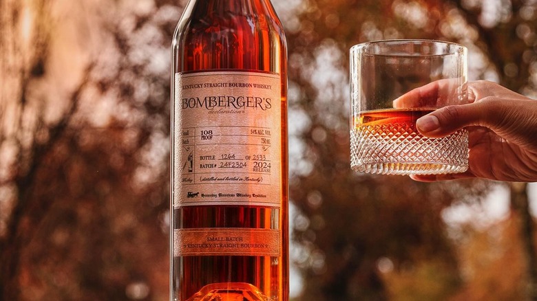A bottle of Bomberger's Declaration in front of a blurred nature setting next to a hand holding up a filled glass