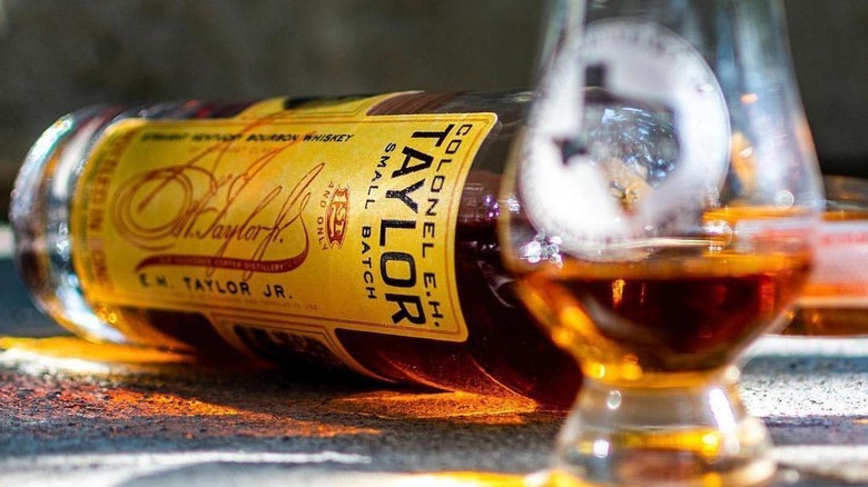 A bottle of Colonel E.H. Taylor, Jr. Small Batch laid on its side with a blurred filled whiskey glass in the foreground