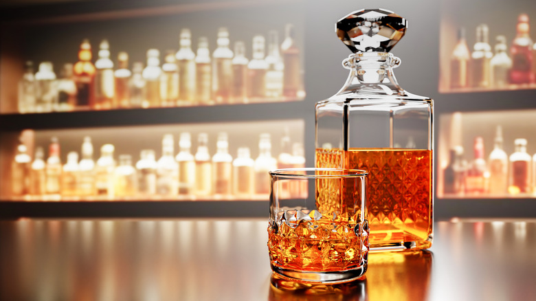 A stylized shot of a brightly lit decanter and filled whiskey glass on a bar with bottles shown in the background