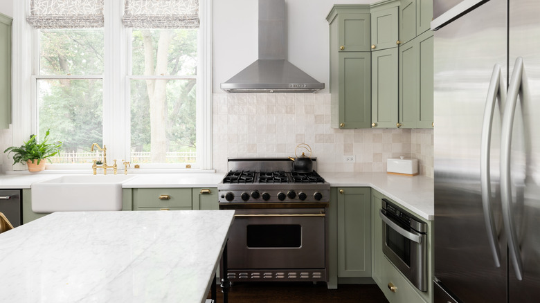 A bright kitchen with sage green cabinets and white walls, tile backsplash, and marble countertops