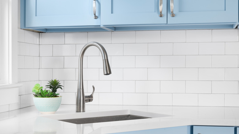 A close up of a silver kitchen sink with a white tile backsplash and light blue cabinets