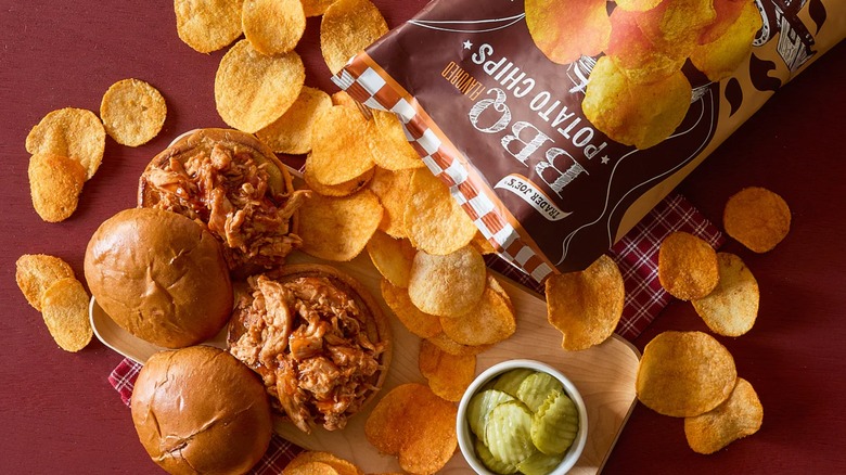 Trader Joe's BBQ Potato chips poured out on a table with two barbecue sandwiches on a wooden board