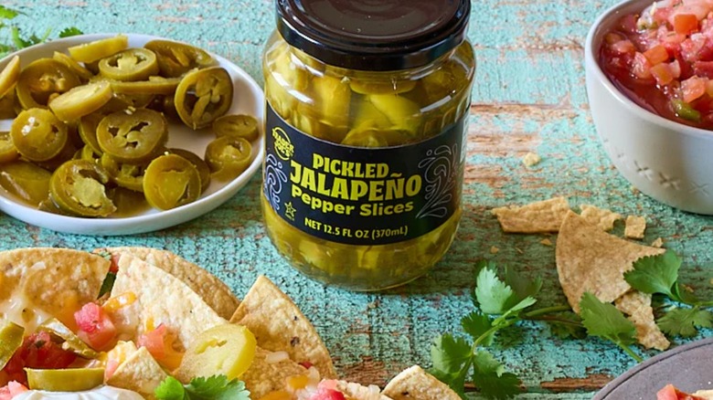 A jar of Trader Joe's Pickled Jalapeño Pepper Slices on a table with a plate of pepper slices, salsa, and nachos