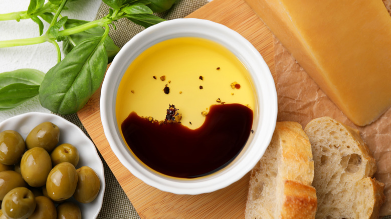 Top view of a sauce bowl filled with olive oil and soy sauce with bread, olives, and fresh basil on the side