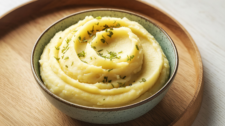 Bowl of creamy mashed potatoes served sprinkled with herbs on a round wooden tray