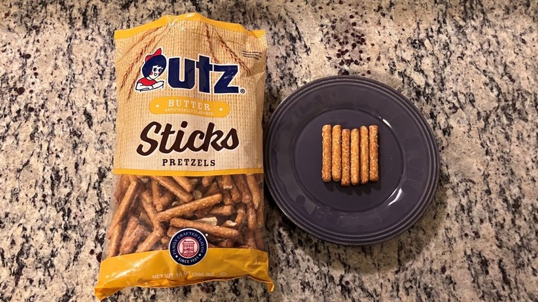 A bag of Utz Butter Pretzel Sticks sits on a countertop next to a purple plate with six pretzels on it