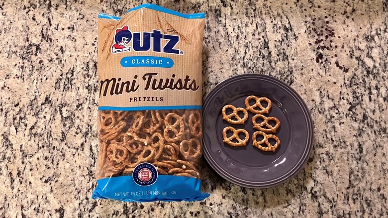A bag of Utz Mini Twists Pretzels sits on a countertop next to a purple plate with five pretzels on it