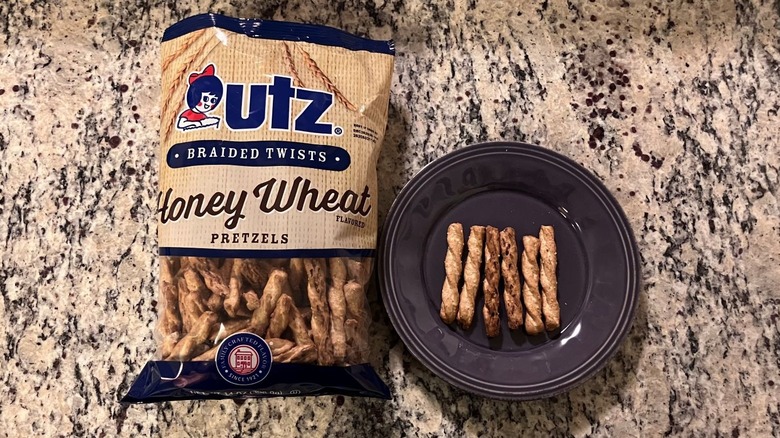 A bag of Utz Braided Twists Pretzels Honey Wheat sits on a countertop next to a purple plate with six pretzels on it