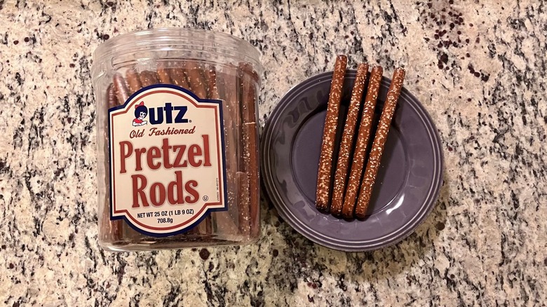 A barrel of Utz Old Fashioned Pretzel Rods sits on a countertop next to a purple plate with four pretzel rods on it
