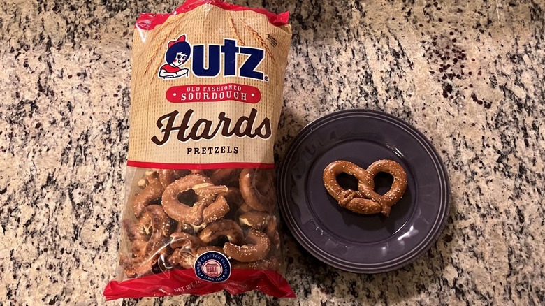 A bag of Utz Old Fashioned Sourdough Hards Pretzels sits on a countertop next to a purple plate with one pretzel on it