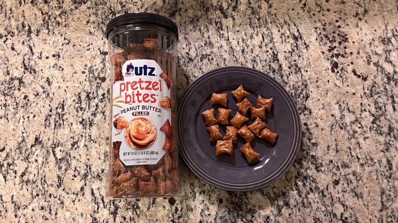 A barrel of Utz Peanut Butter Pretzel Bites sits on a countertop next to a purple plate with fifteen pretzels on it