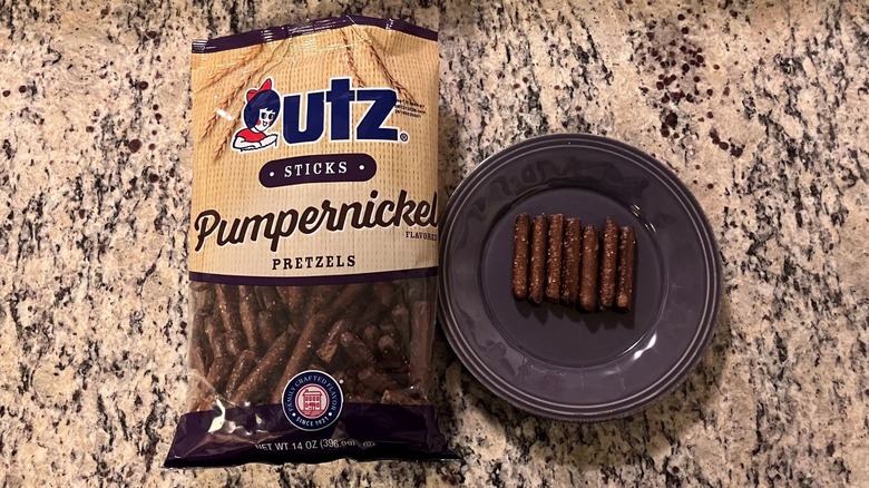 A bag of Utz Sticks Pumpernickel Pretzels sits on a countertop next to a purple plate with seven pretzels on it