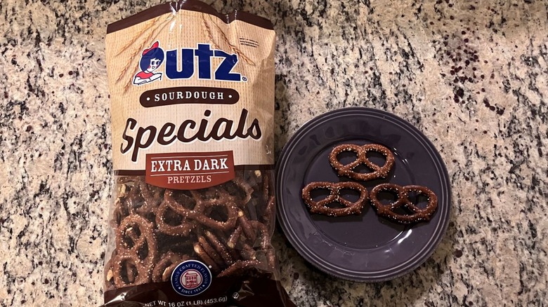 A bag of Utz Sourdough Specials Extra Dark Pretzels sits on a countertop next to a purple plate with three pretzels on it