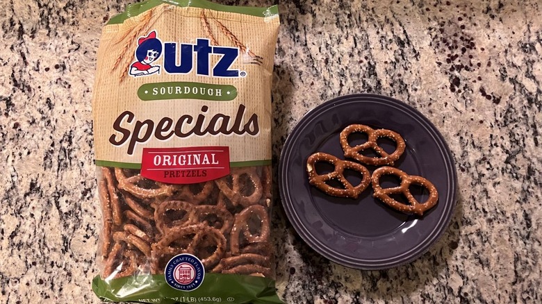A bag of Utz Pretzels Sourdough Specials Original sits on a countertop next to a purple plate with three pretzels on it