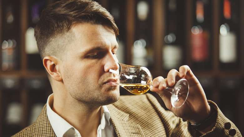 A person drinking whiskey from a tasting glass with blurred bottles in the background