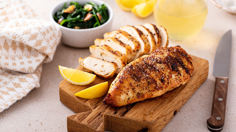 two spice-crusted chicken breasts, one whole and one sliced, shown on a cutting board with lemon wedges, a knife alongside, and a bowl of greens in soft focus to the rear