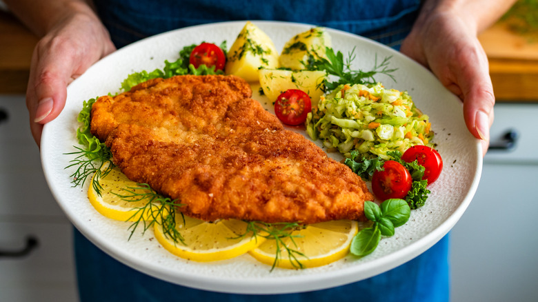 woman holding a white plate with a chicken schnitzel served over lemon slices and fresh herbs, with boiled potatoes, slaw, and a garnish of halved cherry tomatoes