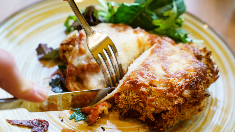 knife and fork cutting into a portion of golden, cheese-topped chicken Parmigiana, served on a yellow plate with a side salad of fresh greens