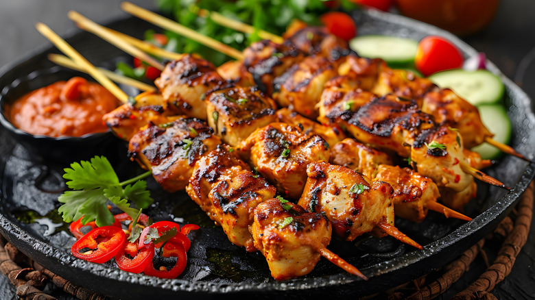 five skewers of grilled chicken satay, shown on a black plate with a small bowl of chunky red sauce, and fresh herb and vegetable garnishes