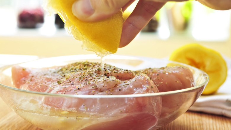 hand squeezing juice from a fresh lemon over two chicken breast halves in a glass bowl filled with marinade