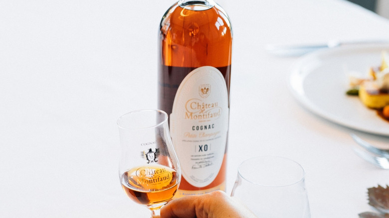 A filled glass of cognac shown to the camera in front of a bottle of Château de Montifaud XO